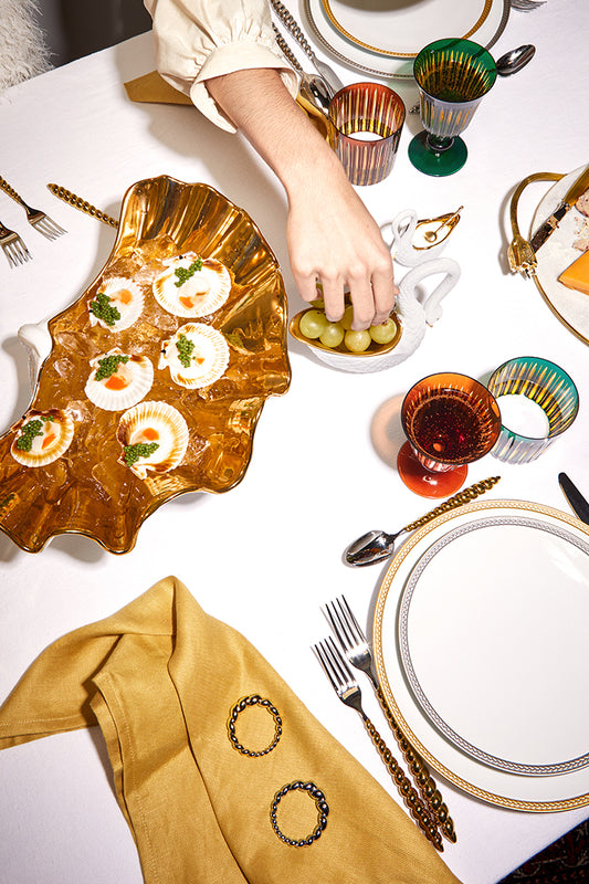 An overhead styled table setting featuring a gold scalloped serving platter with appetizers, a hand placing olives, white plates with gold trim, gold-toned cutlery, and a mustard yellow napkin on a white table.